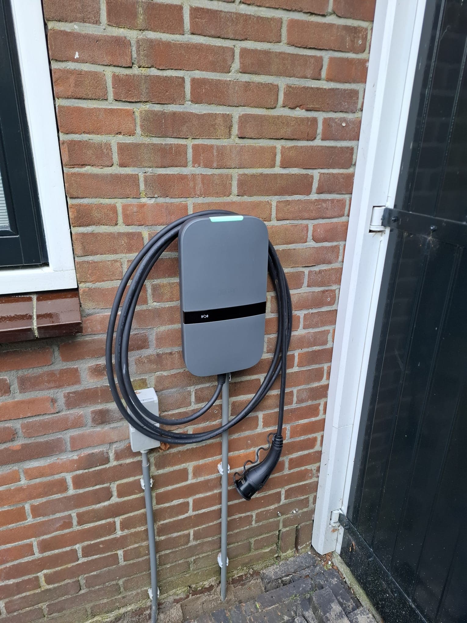 Antracietgrijze Peblar laadpaal aan een bakstenen gevel, met de laadkabel netjes opgehangen.
