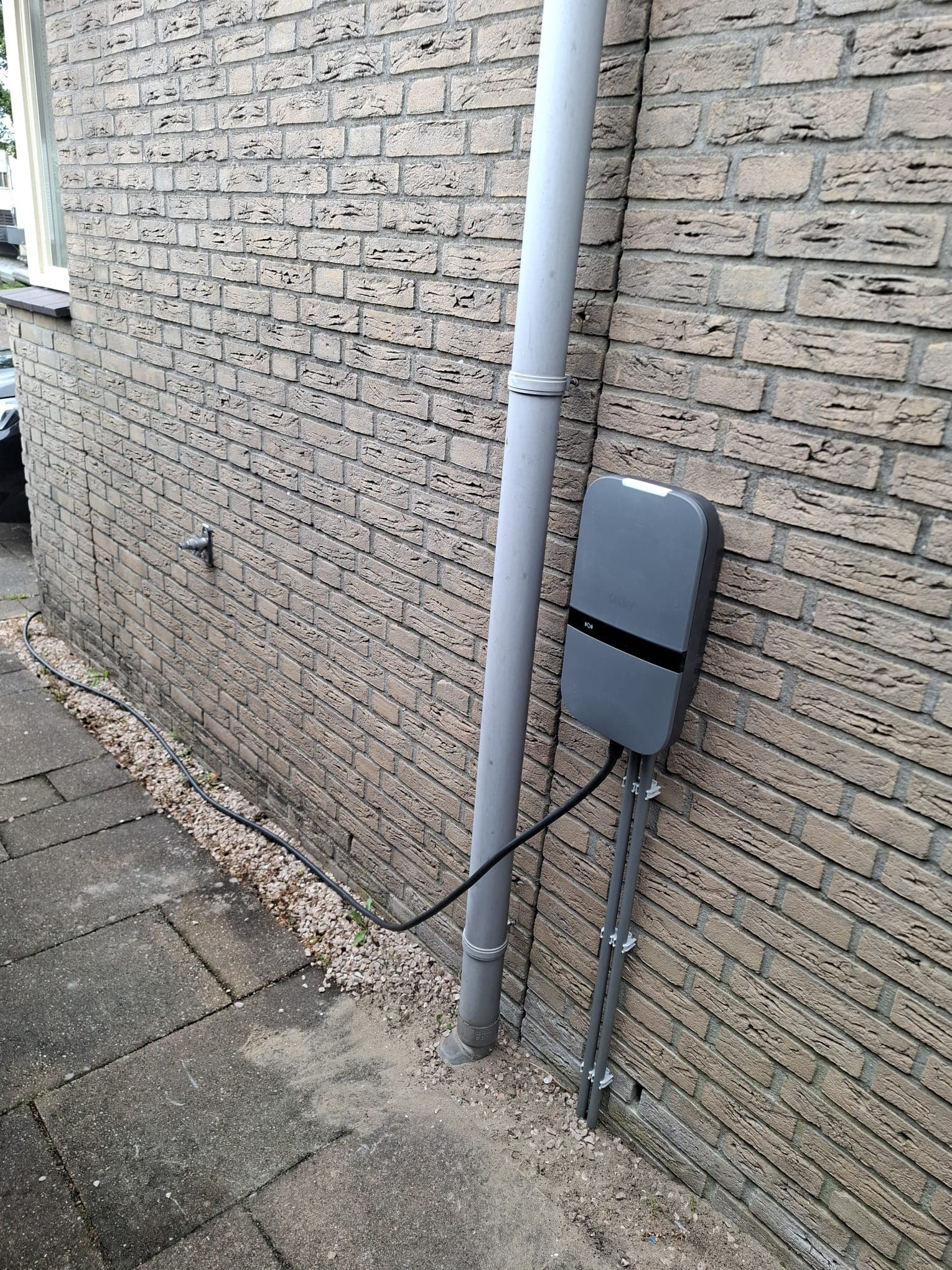 Grijze Peblar laadpaal aan de zijgevel van een woning, met de kabel weggewerkt langs de gevel.
