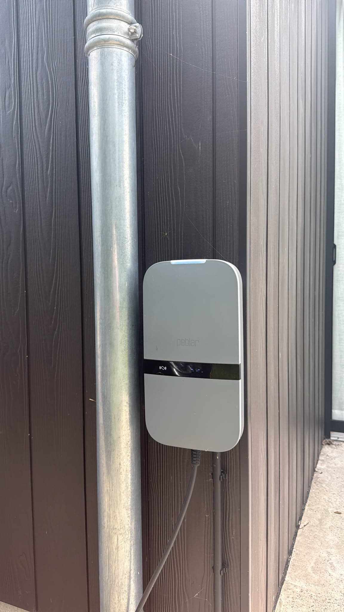 Witte Peblar laadpaal tegen een houten gevelbekleding, strak weggewerkt langs de regenpijp.